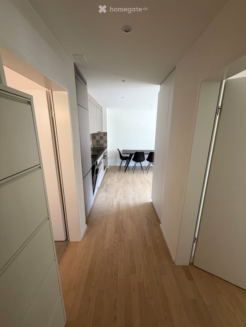 A narrow hallway with light wooden flooring, white walls, and recessed ceiling lighting. The hallway leads to a small kitchen area with cabinets, a countertop, and a dining table with chairs.