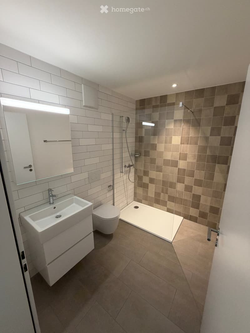 Modern bathroom with white tiles, a sink, toilet, shower, and a large mirror with built-in lighting.