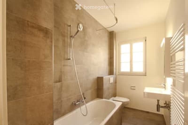 Modern bathroom with a bathtub, toilet, sink, and showerhead. The walls are tiled in a warm, earthy tone, and the floor is covered with a neutral-colored tile.