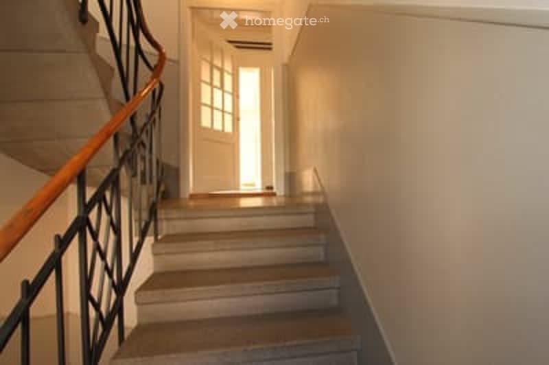 A narrow hallway with a staircase leading upwards, featuring a wooden handrail and a white door at the end.