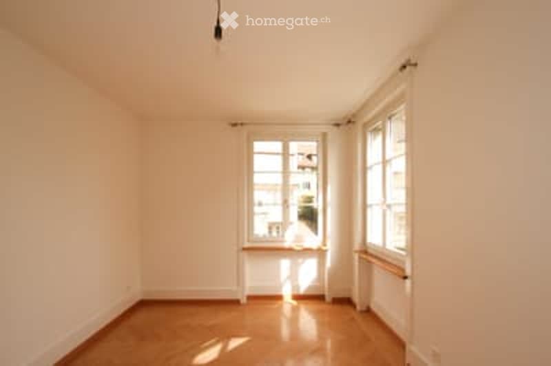 Empty room with wooden flooring, white walls, and large windows with white frames. Natural light streams in, creating a bright and airy atmosphere.