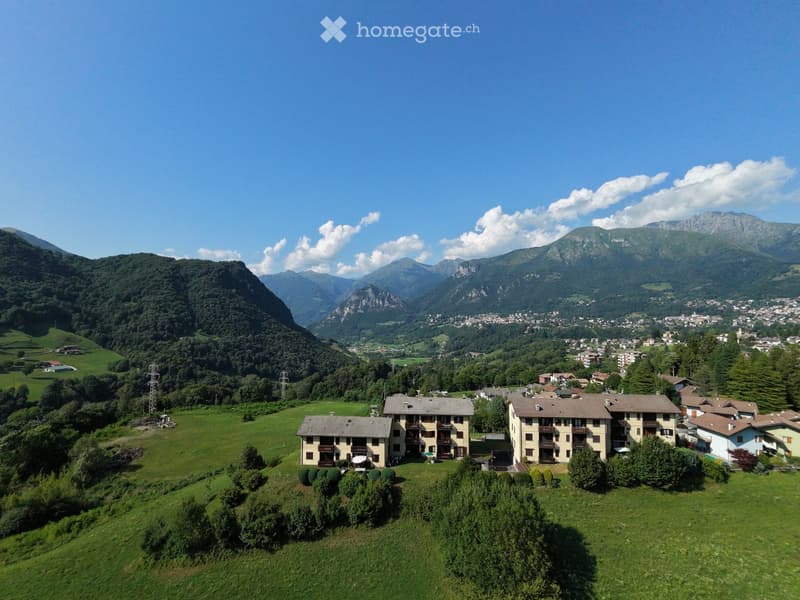 Aerial view of a residential complex with multiple buildings, surrounded by green fields and mountains in the background.