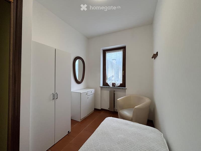 Bedroom with a bed, a white wardrobe, a small cabinet, a round mirror, a window with a curtain, and a beige armchair.