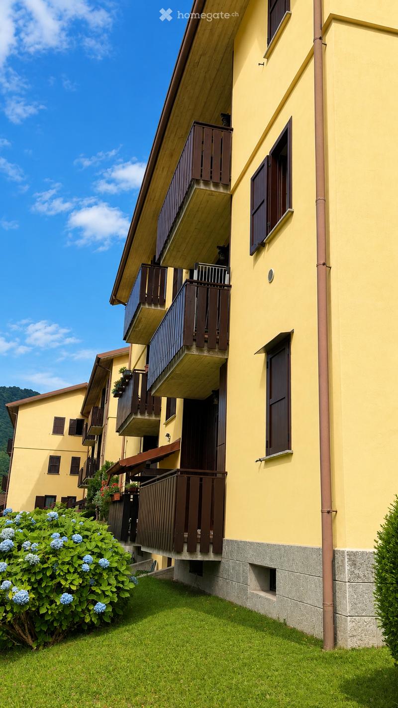 Two-story yellow building with multiple balconies, wooden railings, and shutters. Green lawn and blue flowers in the foreground.