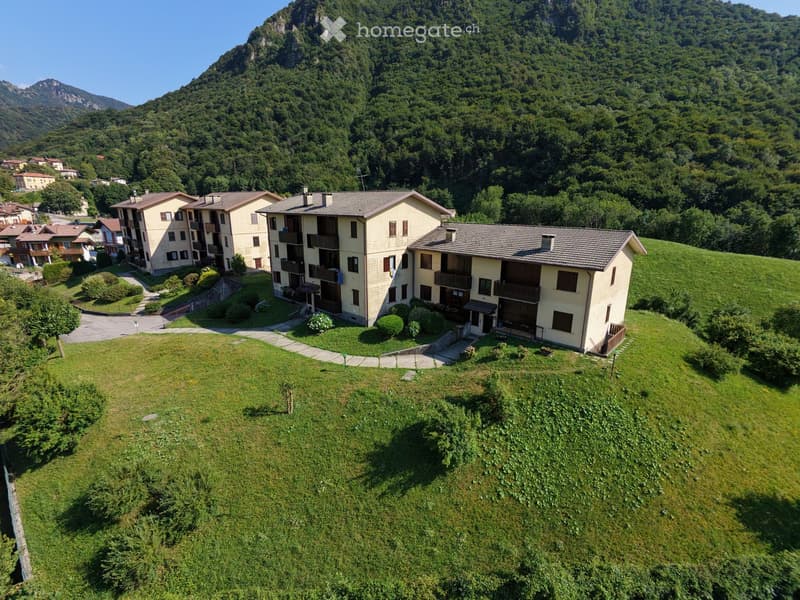 Aerial view of a multi-story residential building complex surrounded by green lawns and trees, with a mountainous backdrop.