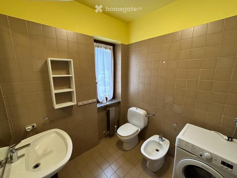 Bathroom with beige tiles, a toilet, bidet, sink, washing machine, and a small shelf on the wall.
