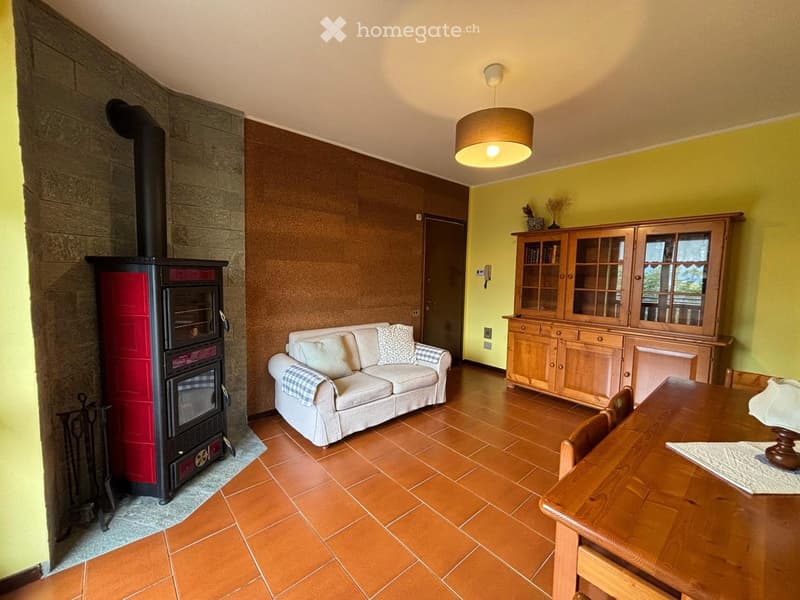 Living room with a tiled floor, a cream-colored sofa with pillows, a wooden dining table with chairs, a wooden cabinet with glass doors, and a red and black wood-burning stove.