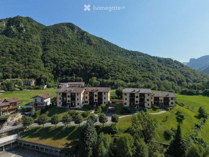 Aerial view of a multi-building complex nestled in a lush green valley with surrounding mountains. The complex includes several residential buildings with balconies and terraces, surrounded by trees and open green spaces.