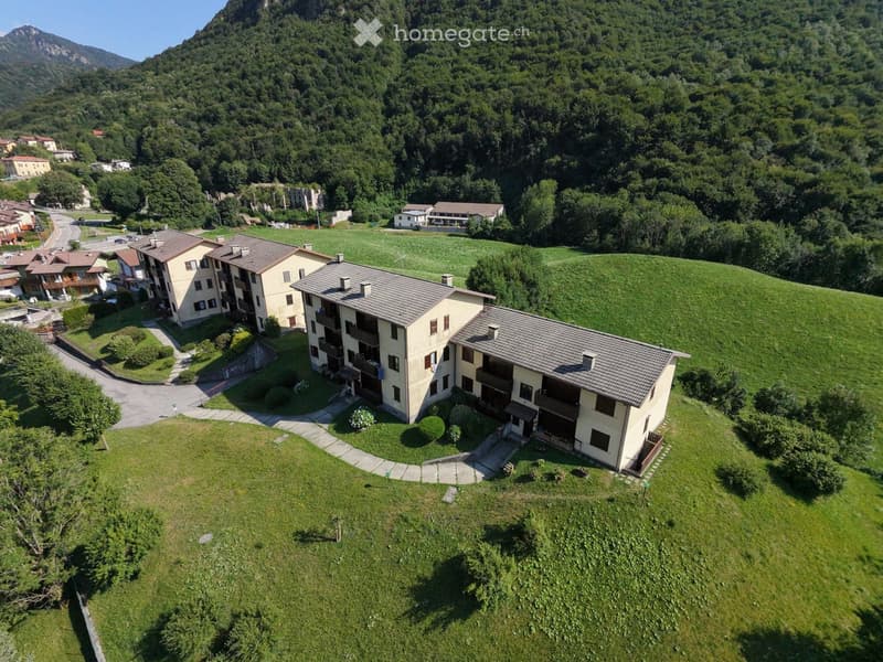 Aerial view of a multi-story residential building complex surrounded by green fields and trees, with a winding road leading to the entrance.
