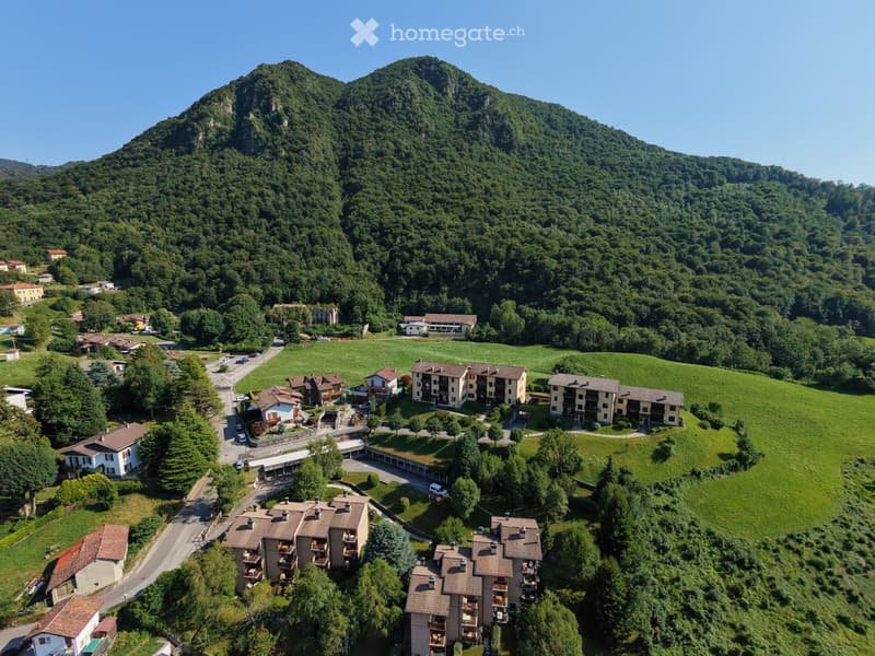 Aerial view of a residential area nestled in a lush green valley surrounded by forested hills and mountains. Multiple houses and apartment buildings are visible, with roads and parking areas connecting them.