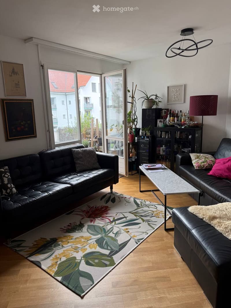 Modern living room with wooden flooring, large windows, black leather sofas, coffee table, floor lamp, bookshelf, and decorative plants.