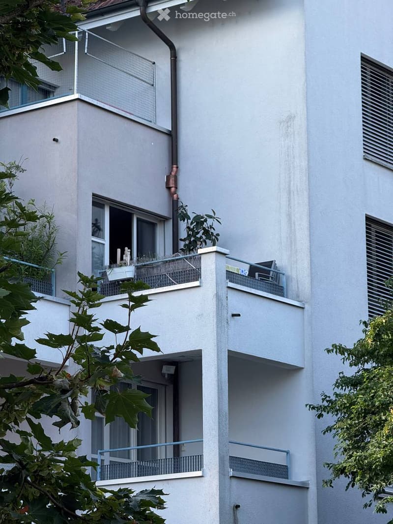 Modern multi-story residential building with white facade, multiple balconies, and greenery visible on the balconies.