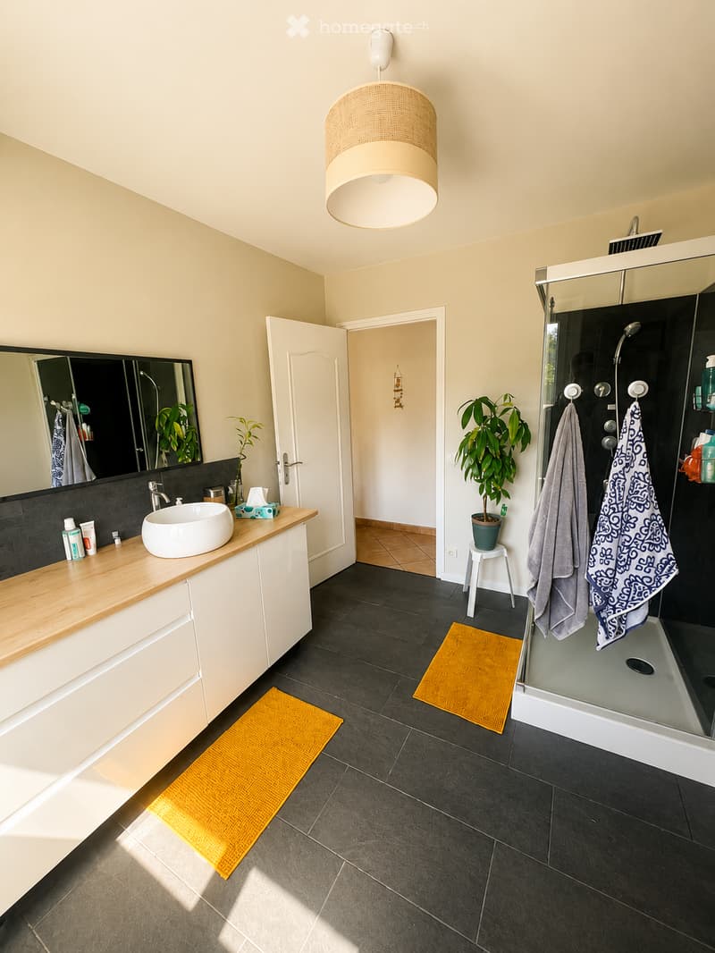 Modern bathroom with a large sink, mirror, and a glass-enclosed shower. Features include a wooden countertop, dark tiled floor, and decorative yellow rugs.