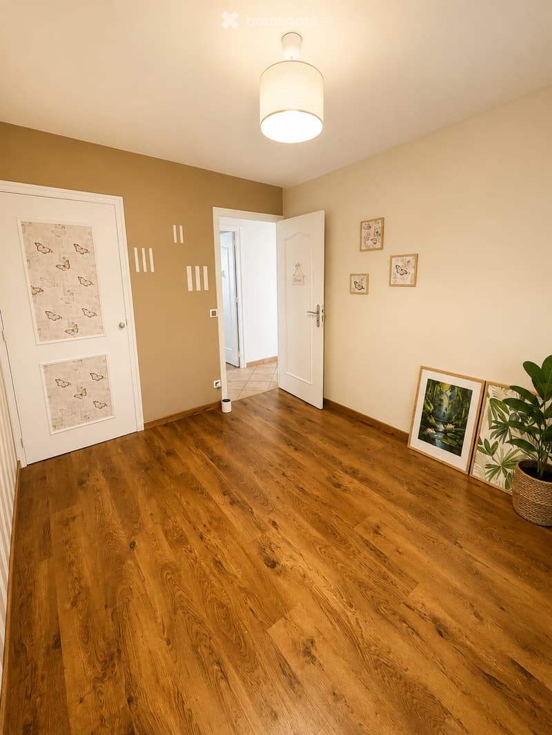 Empty hallway with wooden flooring, white walls, and a pendant light fixture. Features two doors, one with decorative glass panels, and framed artwork on the walls.