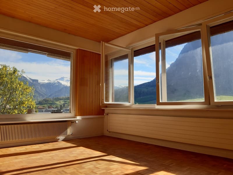 Empty room with wooden flooring, large windows offering a scenic mountain view, and radiators along the walls.