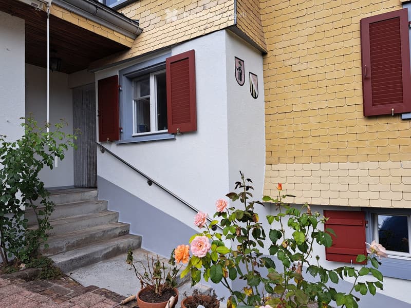 Two-story house with white walls, red shutters, and a yellow roof. Features a small staircase leading to the entrance, surrounded by potted plants and flowers.