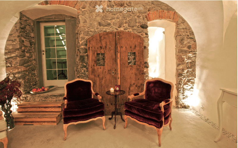 A cozy, rustic room with stone walls and arched doorways. Two plush armchairs in deep purple velvet are positioned facing each other, with a small round table between them holding a candle. A fruit bowl and vase with flowers sit on a wooden bench near the