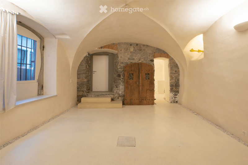 Empty room with arched stone walls, white painted ceiling, and a large wooden door. The floor is light-colored concrete with a small rug in the center. There is a window with white curtains on the left side and a small step leading to another room through