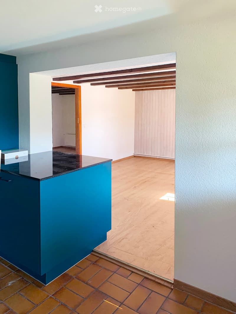 Empty room with tiled floor, wooden ceiling beams, and a small counter area with a blue surface. The room has white walls and an open doorway leading to another space.