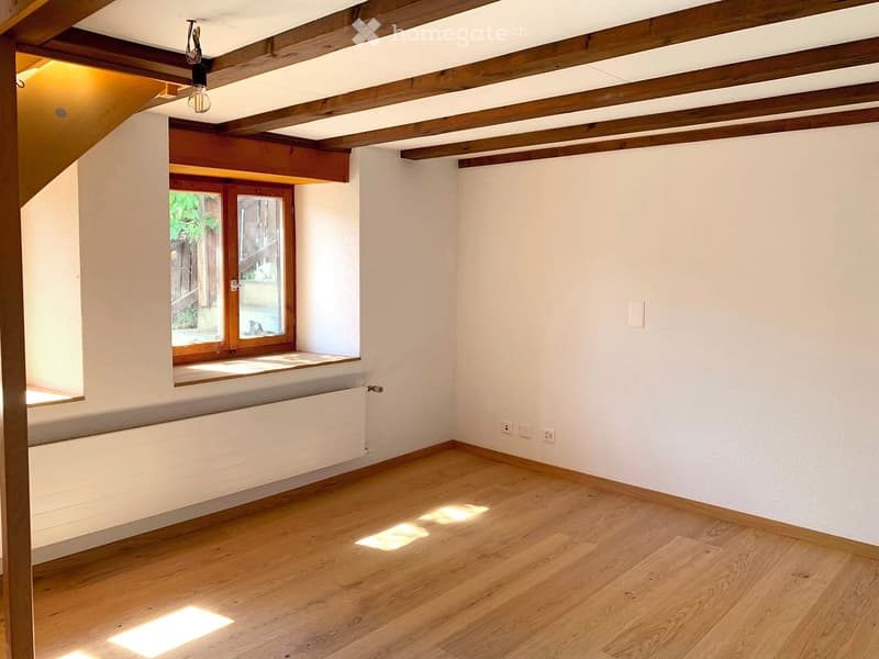 Empty room with wooden flooring, white walls, exposed wooden beams on the ceiling, and a large window with wooden frame and radiator below.