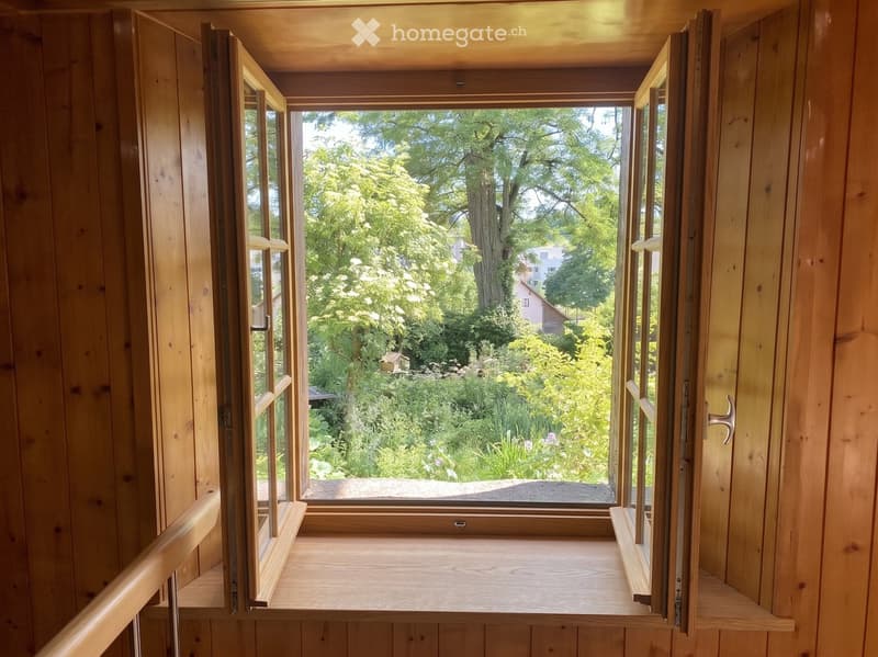 Wooden balcony with open windows, overlooking a lush garden with trees and a small house in the background.