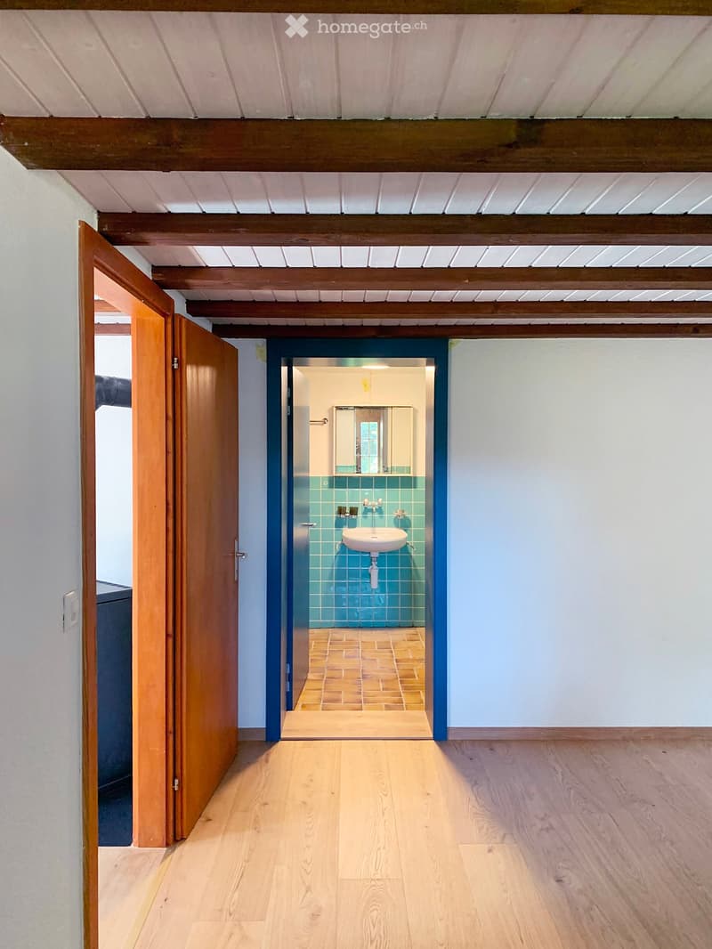 Bright hallway with wooden flooring, white walls, and exposed wooden beams on the ceiling. An open wooden door leads to a small bathroom with blue tiles and a white sink.