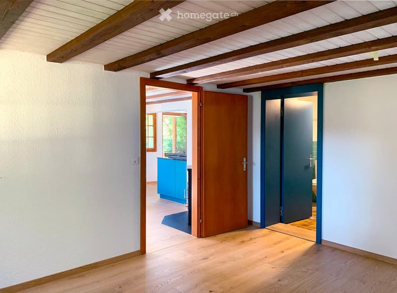 Bright hallway with wooden flooring and exposed wooden beams on the ceiling. Features two open doors leading to other rooms, one with a blue cabinet visible in the background.