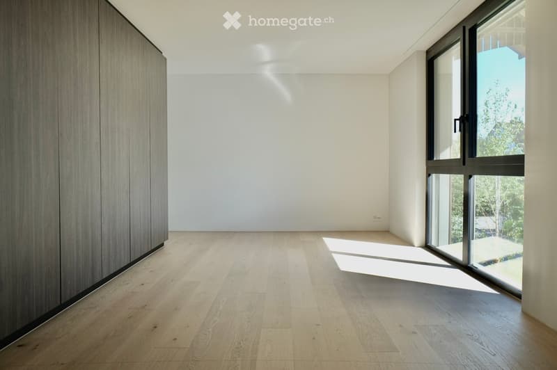 Empty room with light wooden flooring, white walls, and large glass window allowing natural light to enter.