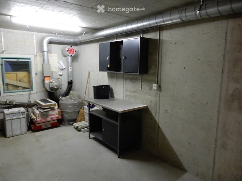 Empty basement room with concrete walls and floor, featuring a workbench, storage items, and a window with a view of stacked materials.