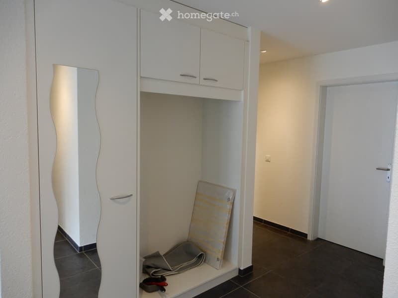 Empty hallway with white walls, dark tiled floor, white cabinet, and a mirror on the left side. A closed door is visible at the end of the hallway.
