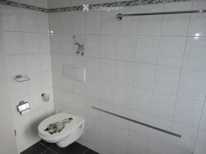 A modern bathroom with white tiled walls, a toilet with a penguin design, a toilet paper holder, a cup holder, and a handrail for support.