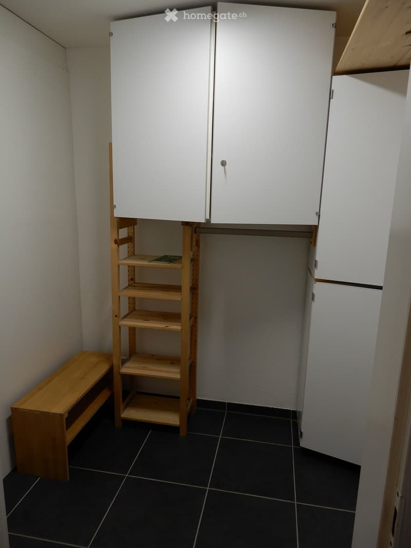 A small, furnished room with a wooden bench, a wooden shelf, and white cabinets.