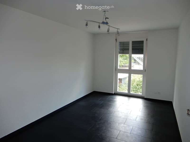 Empty room with white walls, dark tiled floor, and a large window with blinds allowing natural light to enter.