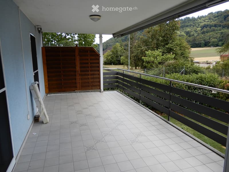 Balcony with tiled floor, metal railing, and wooden gate leading to a grassy area with trees and a field in the background.