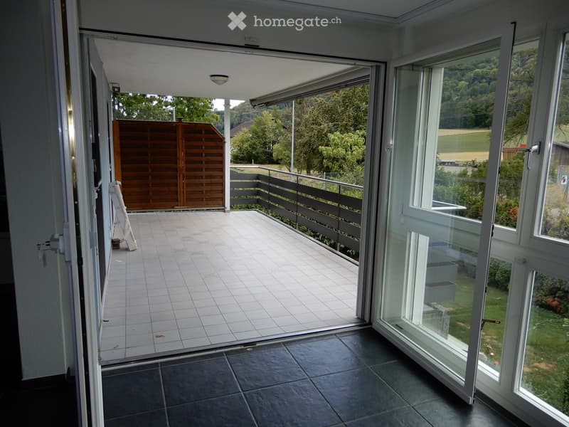 Open sliding glass doors lead to a tiled balcony with a wooden gate and metal railing, overlooking a green landscape.