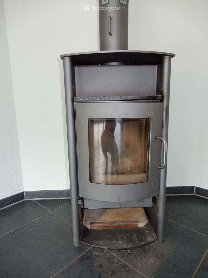 Modern metal wood-burning stove with glass front and ash pan, placed on tiled floor.