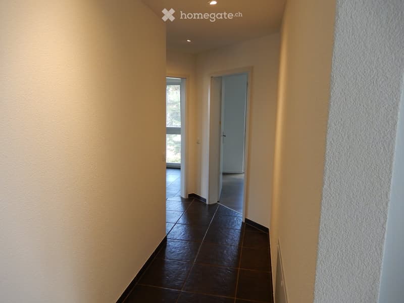 A narrow hallway with dark tiled flooring and light-colored walls, featuring two doors and a view of an adjacent room with a window.