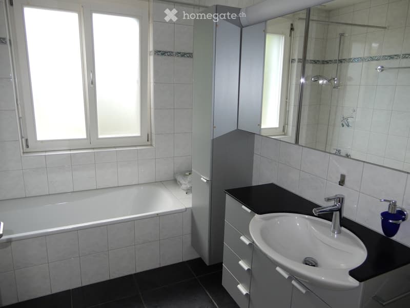 Modern bathroom with a bathtub, sink, mirror, and cabinet. The room features tiled walls and a large window providing natural light.