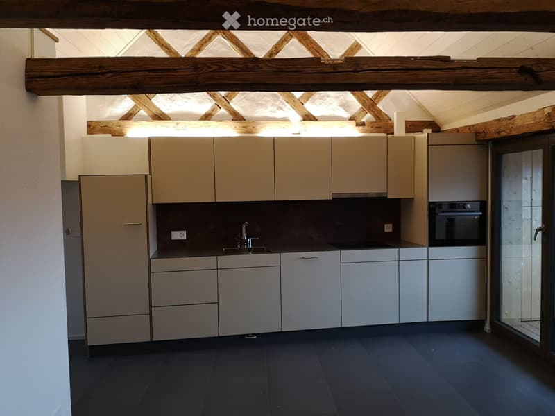 Modern kitchen with light-colored cabinets, stainless steel sink, and built-in oven. Exposed wooden beams on the ceiling and dark tiled floor.
