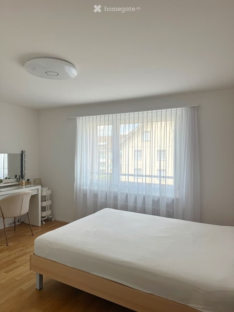 Modern bedroom with a white bed, wooden floor, large window with sheer curtains, vanity table with mirror, and a chair.