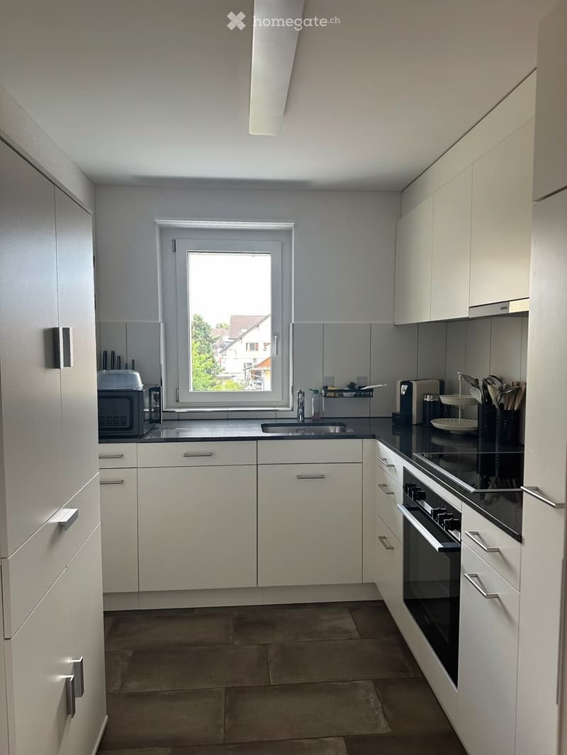 Modern kitchen with white cabinets, stainless steel appliances, and a large window offering a view of the outside. The space includes a sink, oven, microwave, and various kitchen utensils.
