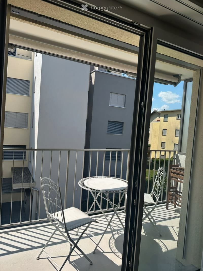 Balcony with a white metal table and two matching chairs, overlooking neighboring buildings.