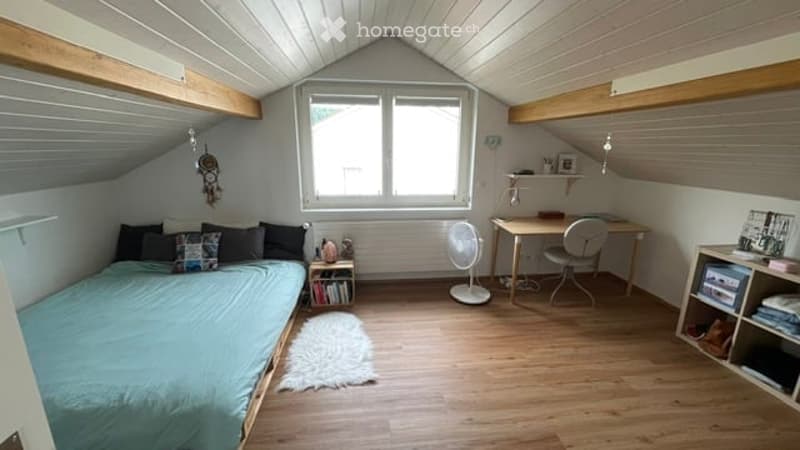 Cozy attic bedroom with slanted wooden ceilings, large window, bed with pillows, desk with chair, fan, shelf, and small rug.