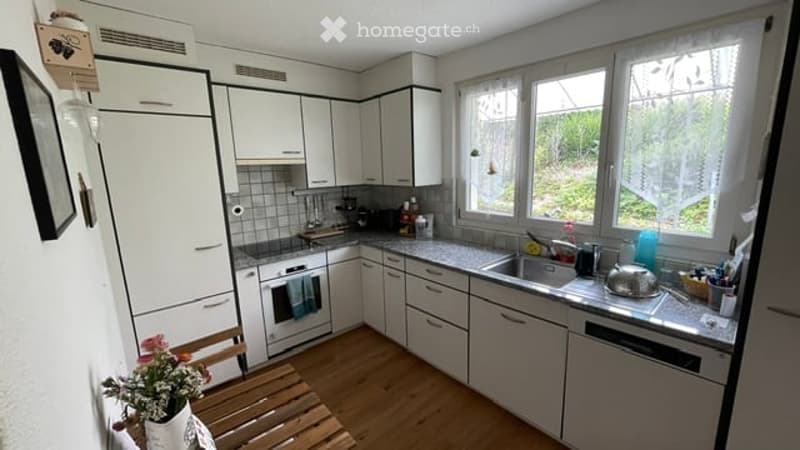 Modern kitchen with white cabinets, stainless steel appliances, and a wooden floor. Features include a refrigerator, oven, sink, dishwasher, and countertop with a teapot and flower pot.