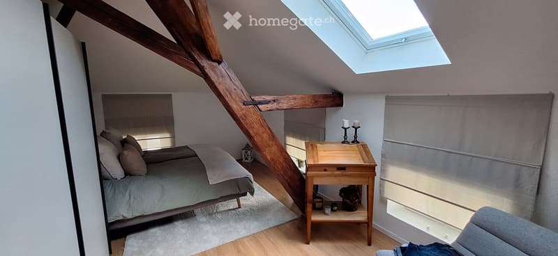 Cozy attic bedroom with wooden beams, skylight, bed with pillows and blanket, nightstand with candles and small plant, and a gray armchair.