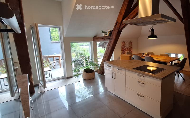 Modern kitchen with white cabinets, a central island with an induction cooktop, and a dining area with a wooden table and chairs. Large windows and sliding doors lead to a patio with outdoor furniture and plants.