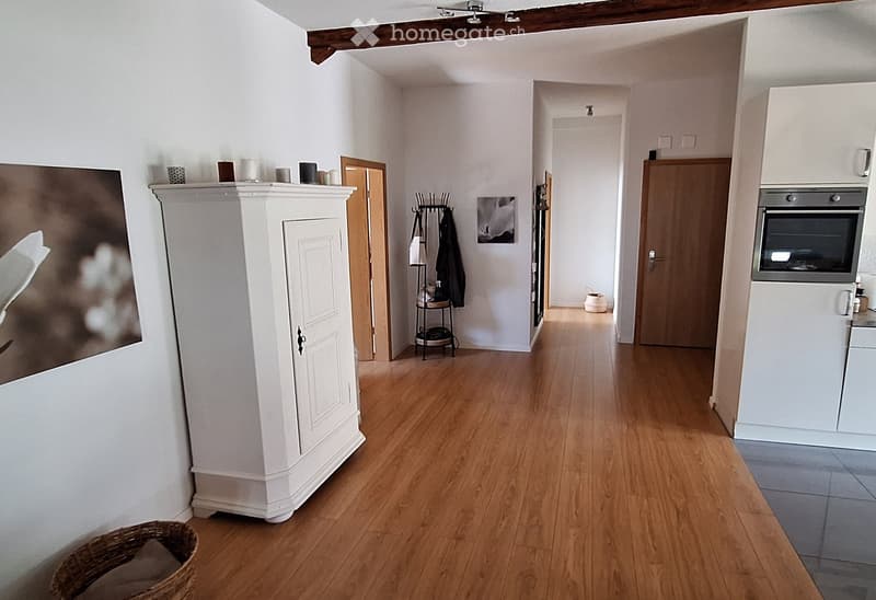 A well-lit hallway with wooden flooring, white walls, and several doors leading to different rooms. There is a white cabinet with decorative items on top, a coat rack with coats, and a small basket on the floor.