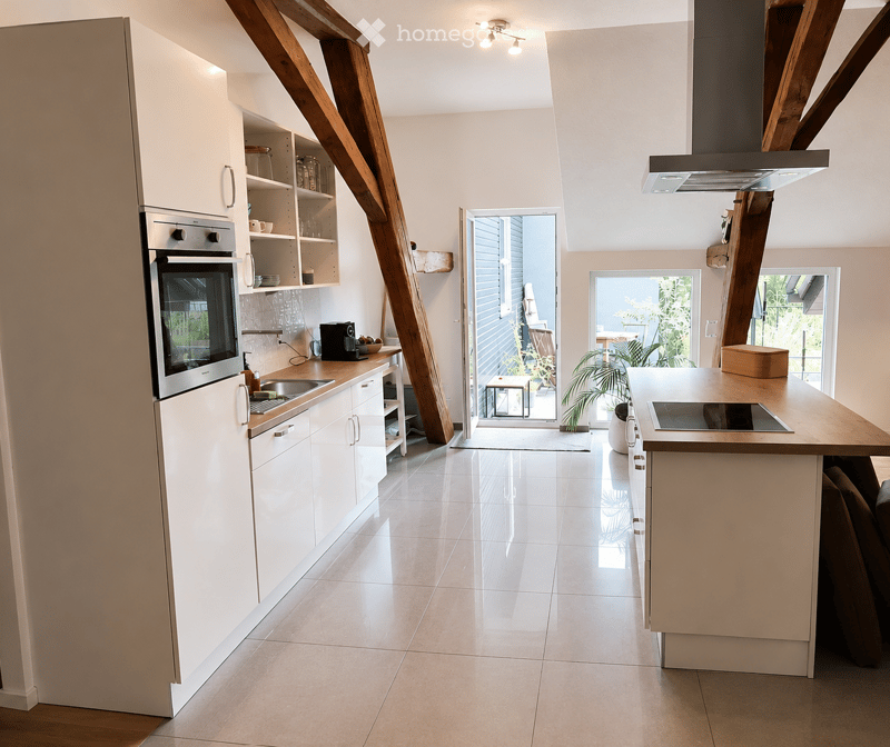 Modern kitchen with white cabinets, stainless steel appliances, and a central island with an induction stove. The space features a sleek design with wooden beams and large windows leading to a balcony.