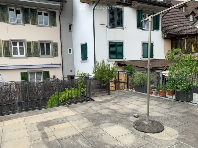 Courtyard with stone pavement, metal fence, potted plants, and surrounding buildings with green shutters.