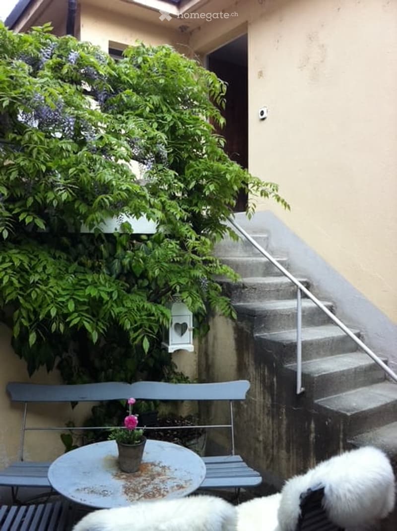A small outdoor area with a bench, a round table, and a flower pot, surrounded by greenery and a staircase leading up.